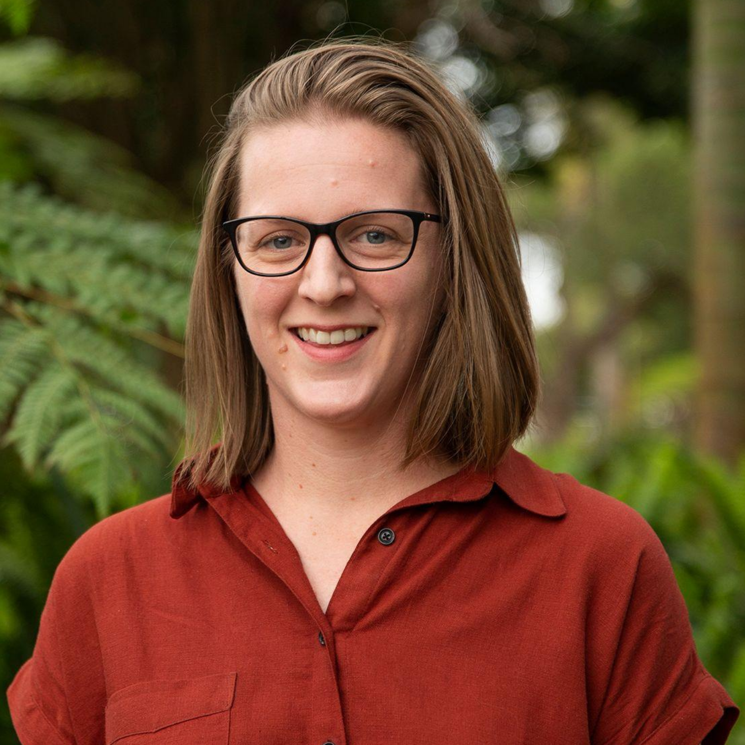 Emma smiles directly at the camera. She has shirt brown hair, glasses and is wearing a red button up shirt