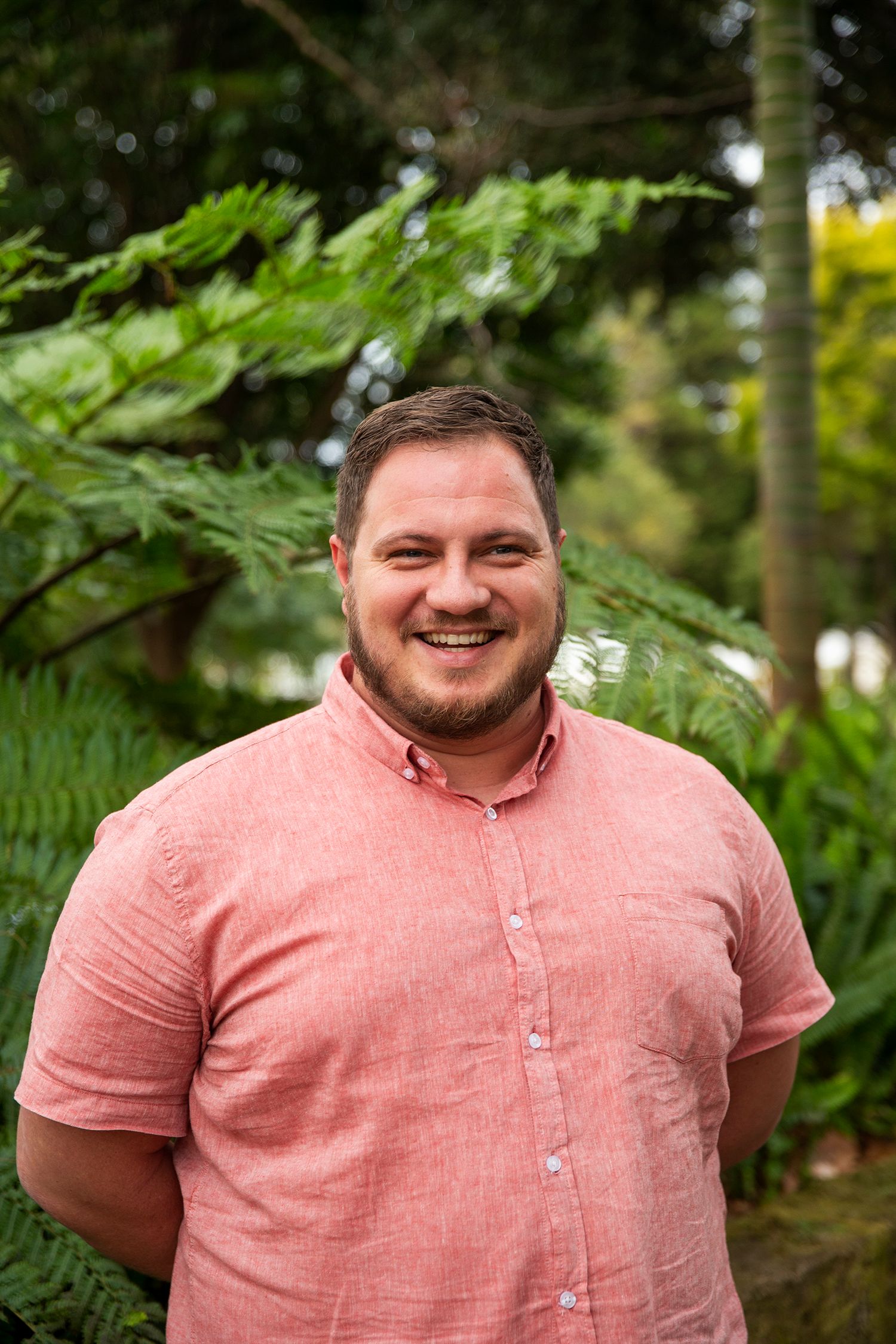 SP smiles directly at the camera. He is wearing a button up coral coloured shirt and has short brown hair and light green eyes with a tanned complexion.
