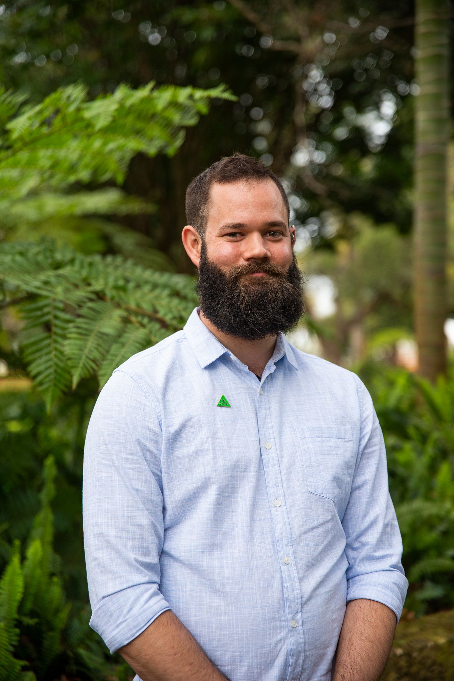 Charles looks directly at the camera, smiling. he is tall, with a big black beard and is wearing a light blue collared shirt.