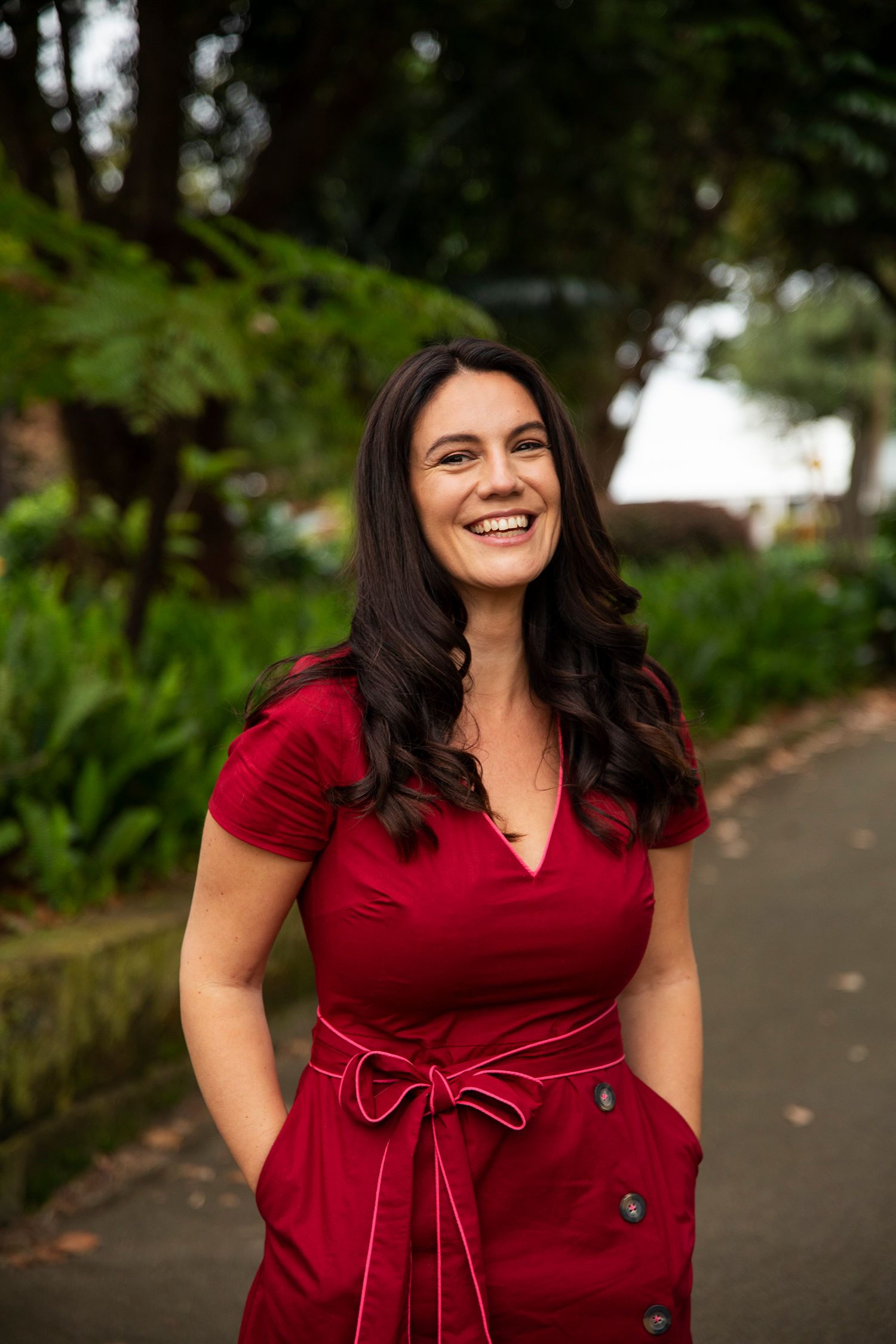 Fran smiles at the camera almost laughing. She has long black hair, a tanned complexion and is wearing a v neck red dress