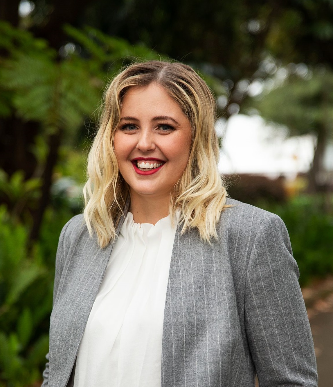 Tamara Alderdice looks directly at camera, smiling. Tamara has short blonde wavey hair, blue eyes, fair skin and wears a greay pin stripe suit with a white blouse underneath. 