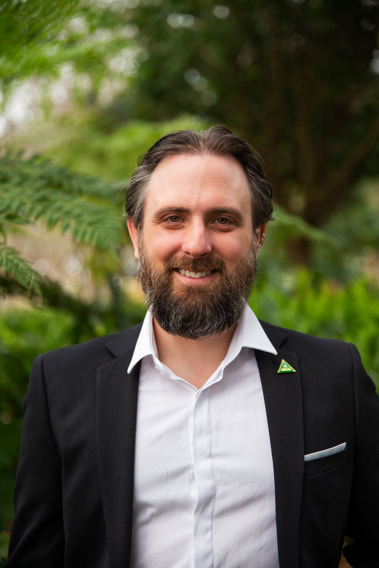 Tim Young looks directly at the camera smiling. he is wearing a suit, has combed back hair and a beard.